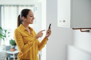 Smiling woman adjusting settings on a wall-mounted boiler using her smartphone at home