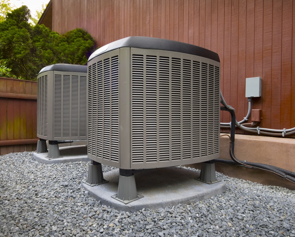 Two outdoor air conditioning units positioned on gravel, beside a wooden fence, with electrical connections visible on the wall