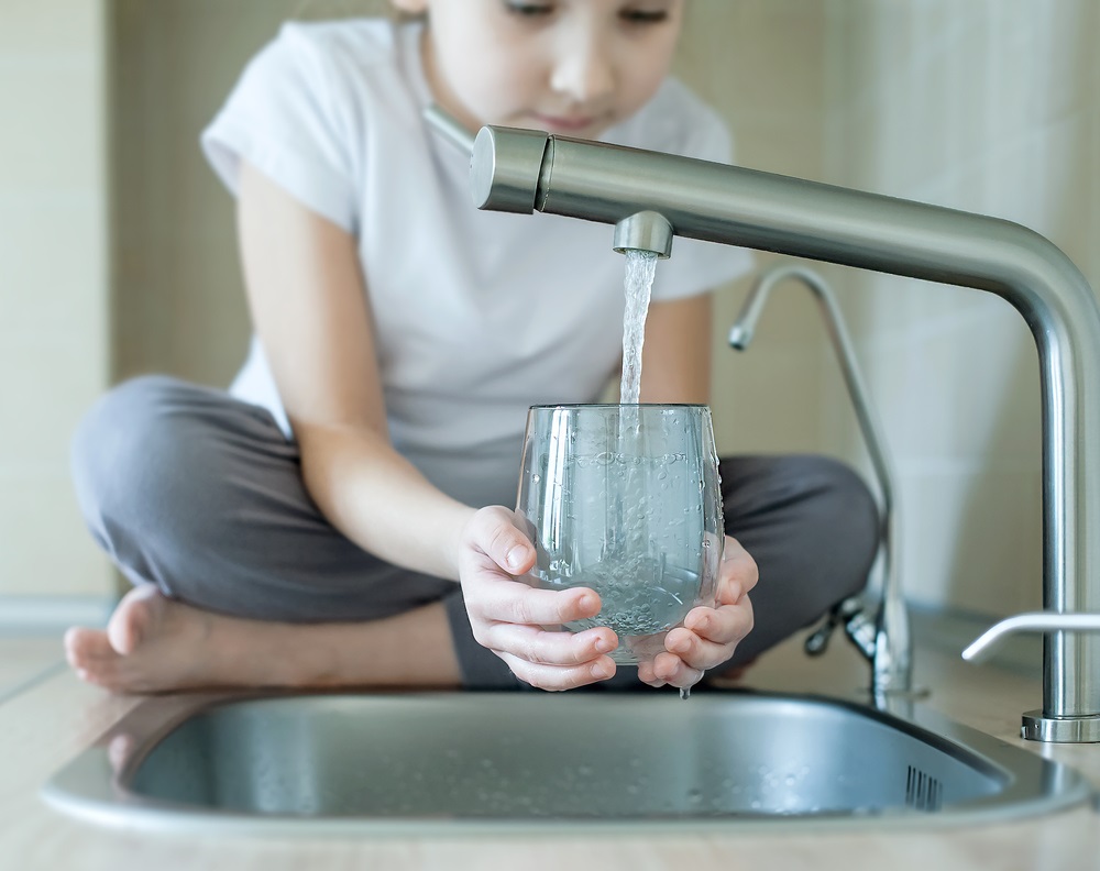person sitting cross-legged by a sink, holding a glass under a flowing faucet, filling it with water.
